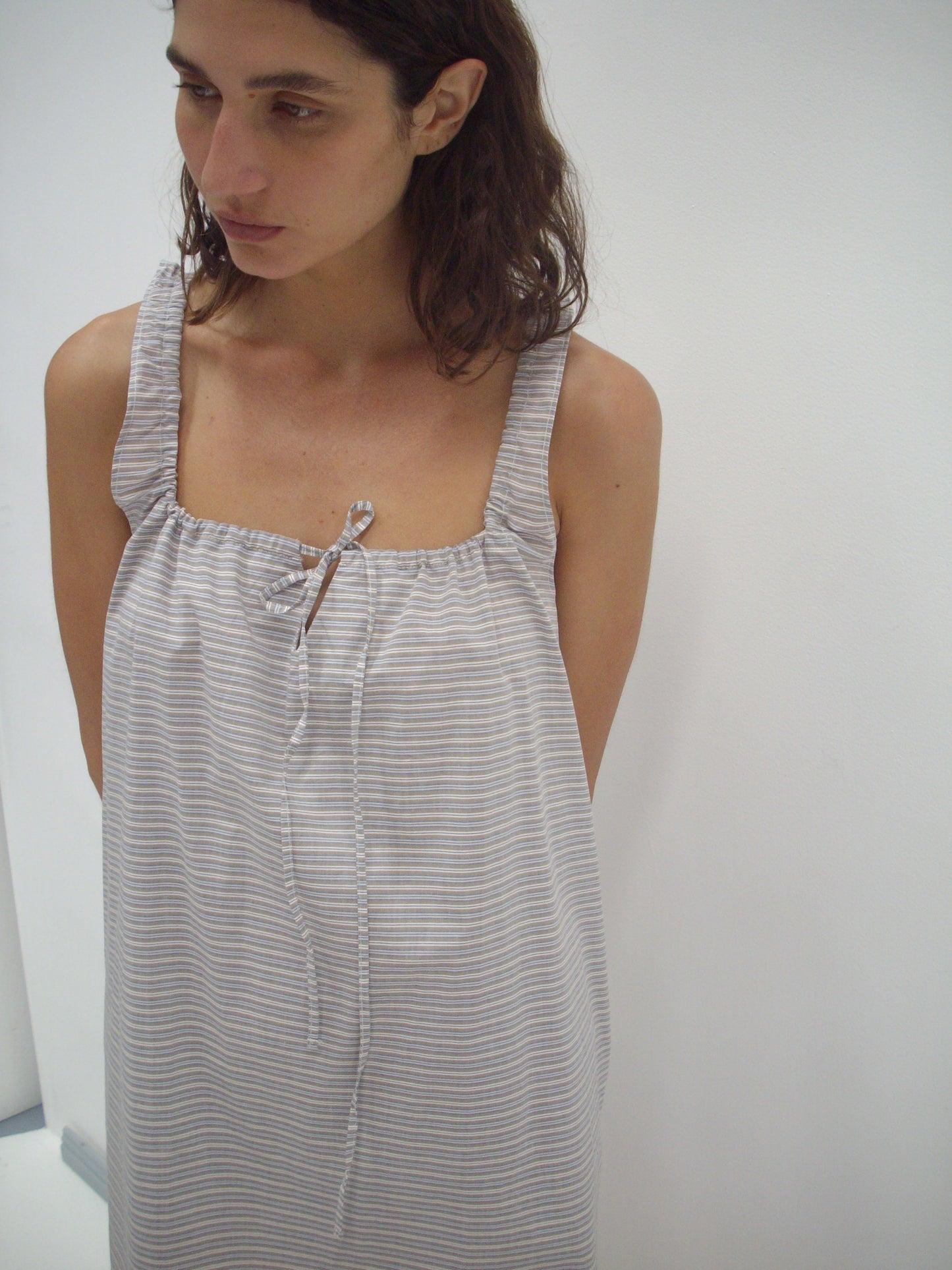 A woman with wavy brown hair wears The Bow Dress - Horizontal Stripe, a loose, sleeveless organic cotton dress in light gray and white stripes with a gathered neckline and string tie. She stands against a plain white background, looking slightly downward.
