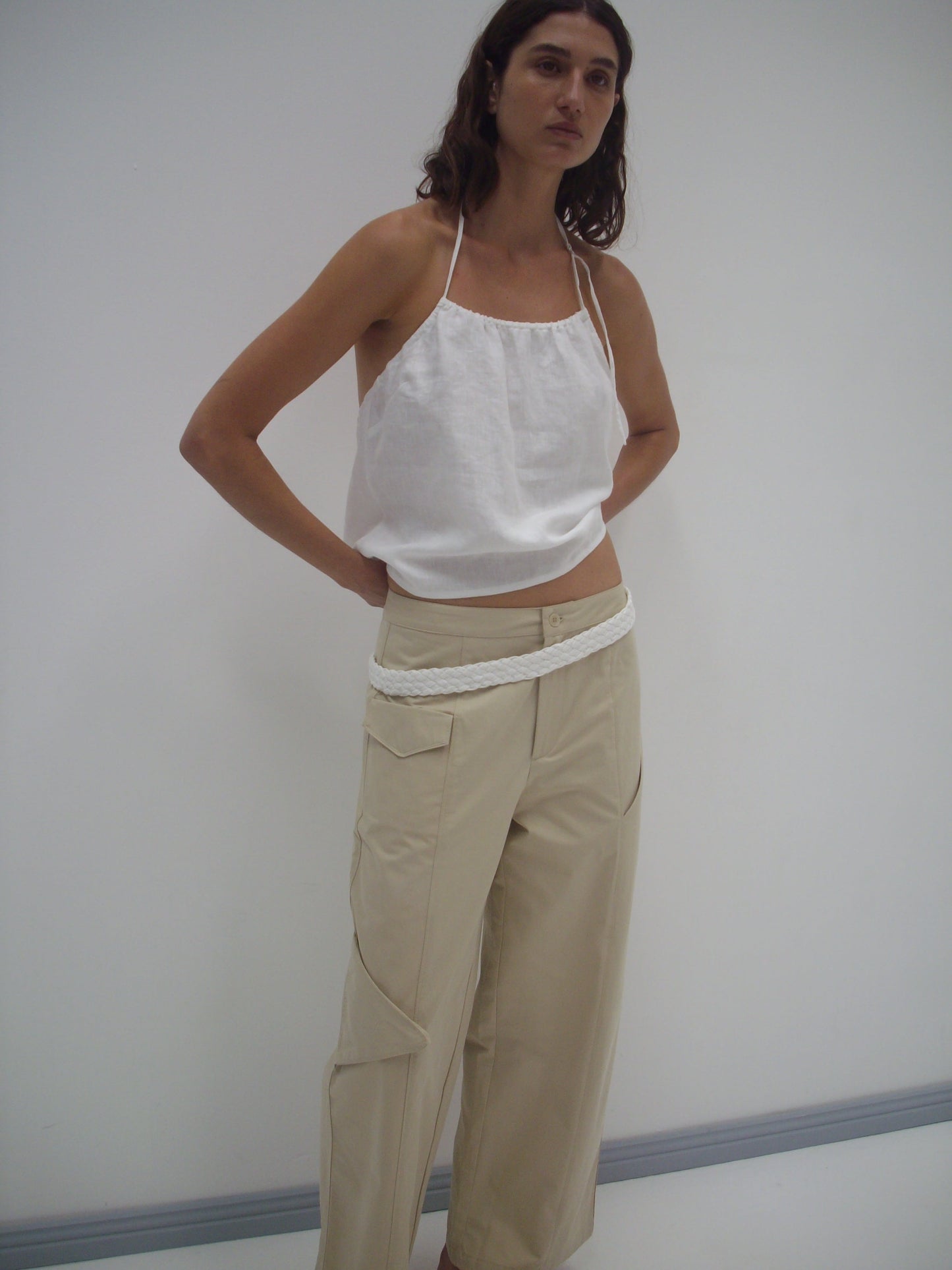 A woman stands against a white wall, wearing a Deiji Studios white sleeveless top and Pocket Pant - Stone—loose beige organic cotton twill pants with cargo pockets and a white braided belt—her hands tucked behind her back.