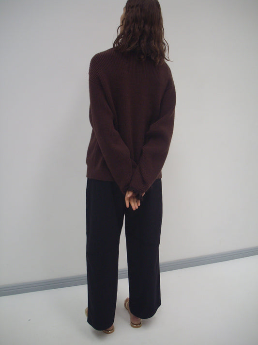 A person with wavy brown hair faces a blank wall, wearing a loose Flounce Cardigan - Burgundy from Deiji Studios, black wide-leg pants, and brown sandals. Their hands are clasped behind their back.