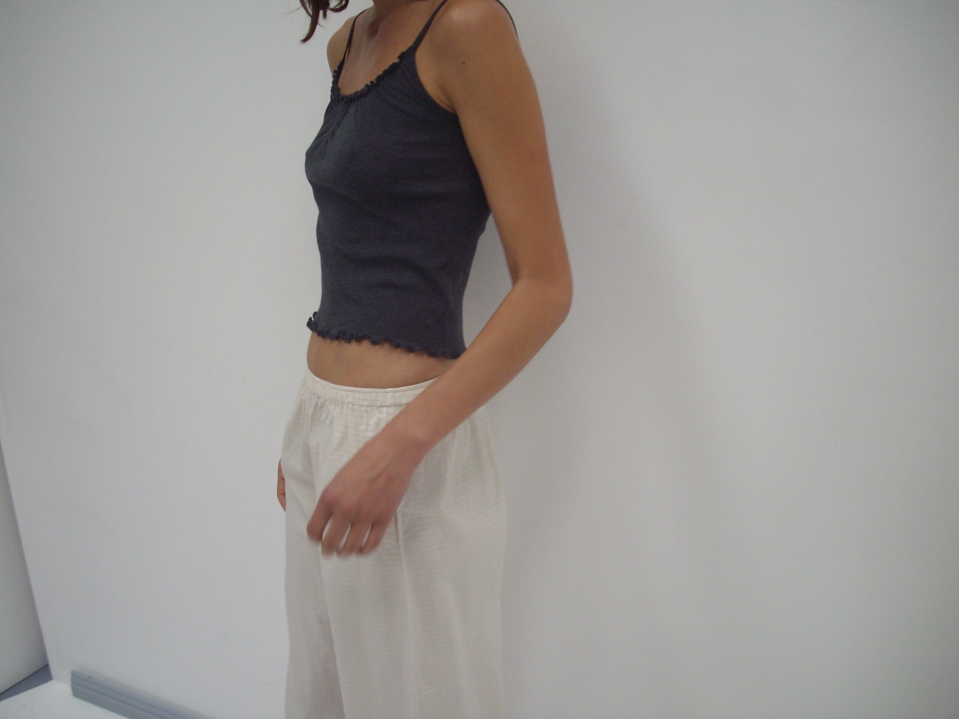 A person stands against a plain white wall, wearing a dark sleeveless top and Deiji Studios' Ease Trouser - Bone Check in organic cotton. Their head is partially out of the frame.