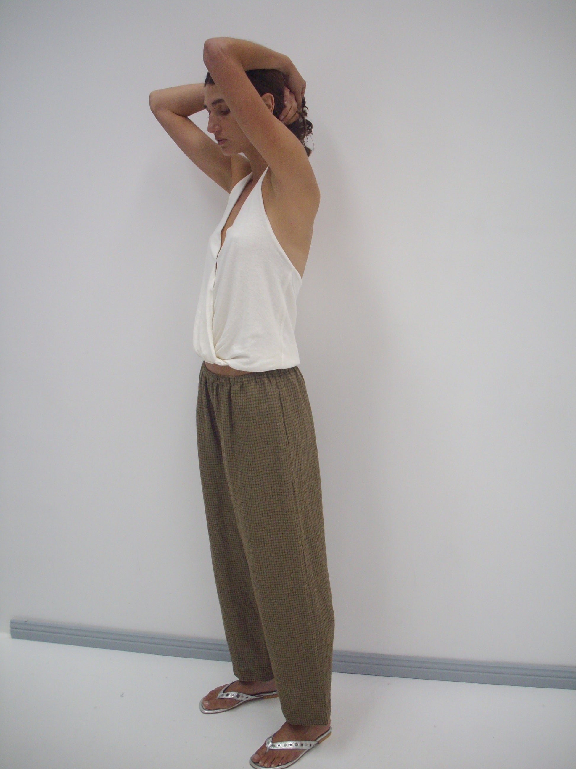A person stands in a minimalist room, wearing a white sleeveless top, the Linen Ease Trouser - Pine Check, and flip-flops. They have their arms raised with hands behind their head, looking relaxed.