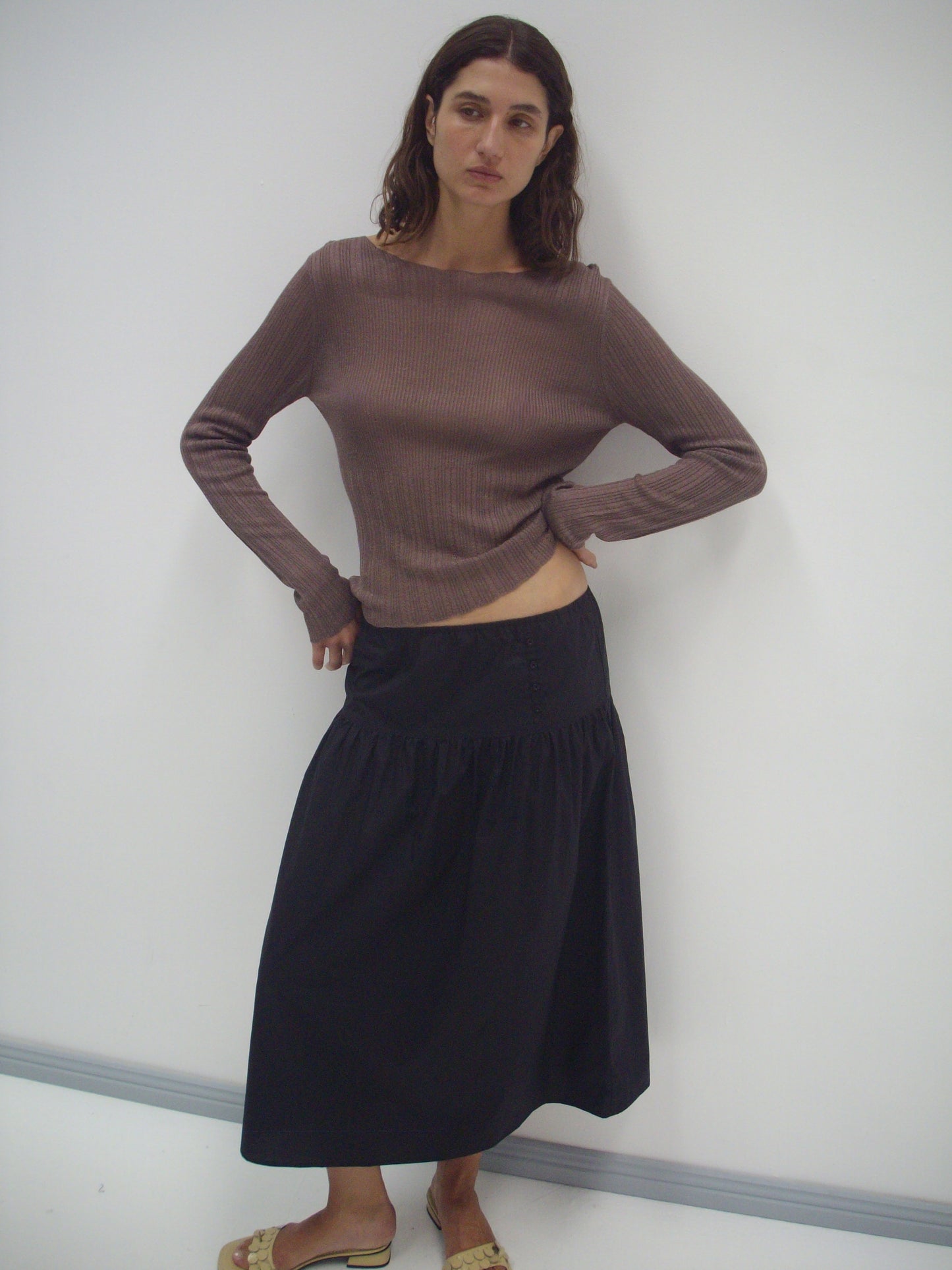 A woman with wavy brown hair stands against a white wall, wearing a long-sleeved brown ribbed top and the Button Skirt - Black. She poses with one hand on her hip and completes the look with tan sandals.