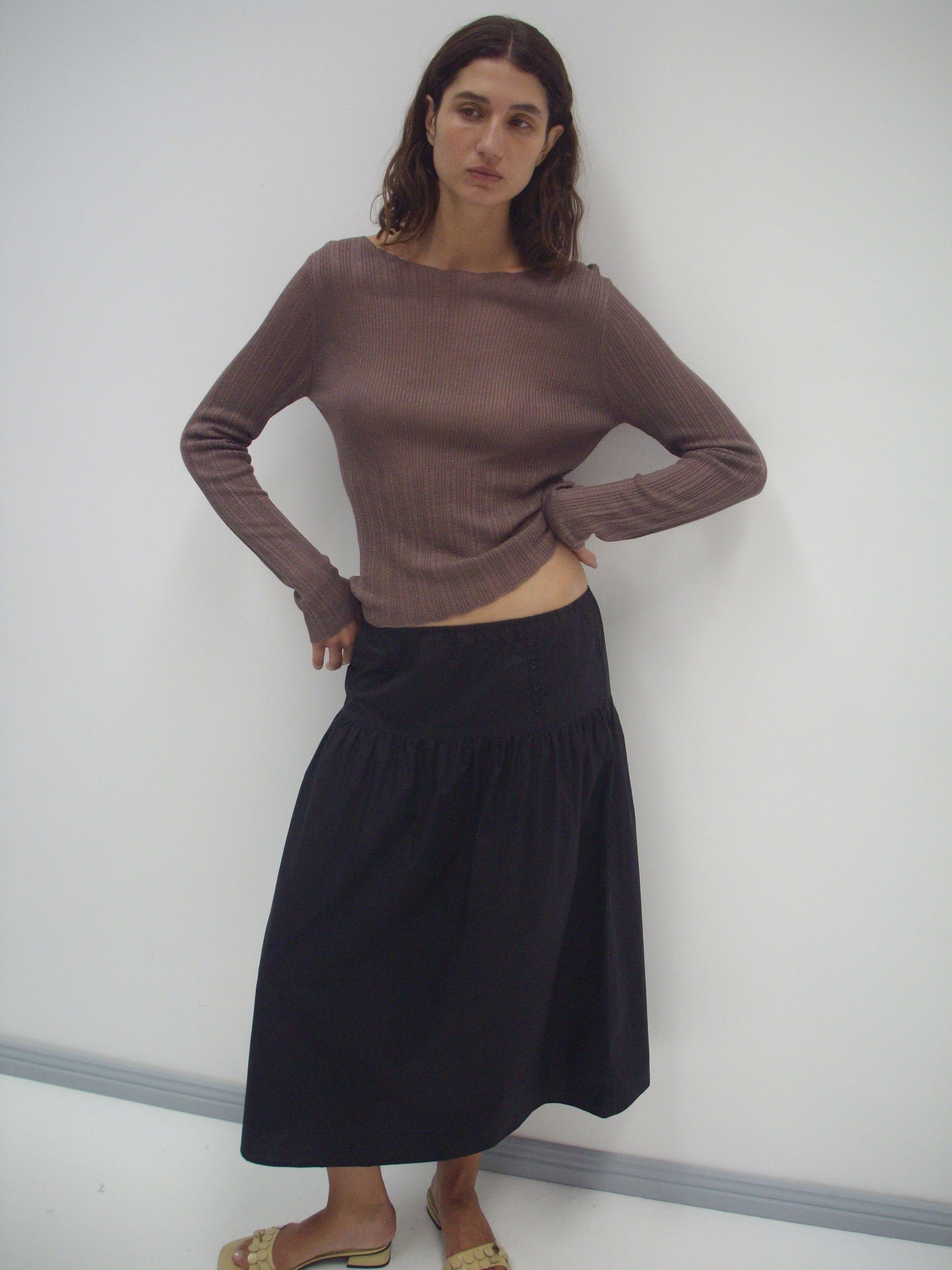 A woman with wavy brown hair stands against a white wall, wearing a long-sleeved brown ribbed top and the Button Skirt - Black. She poses with one hand on her hip and completes the look with tan sandals.