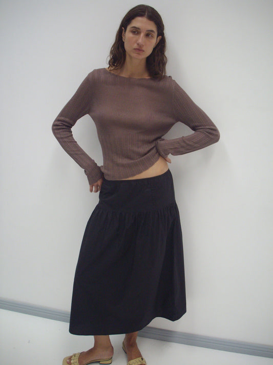A woman with wavy brown hair stands against a white wall, wearing a long-sleeved brown ribbed top and the Button Skirt - Black. She poses with one hand on her hip and completes the look with tan sandals.