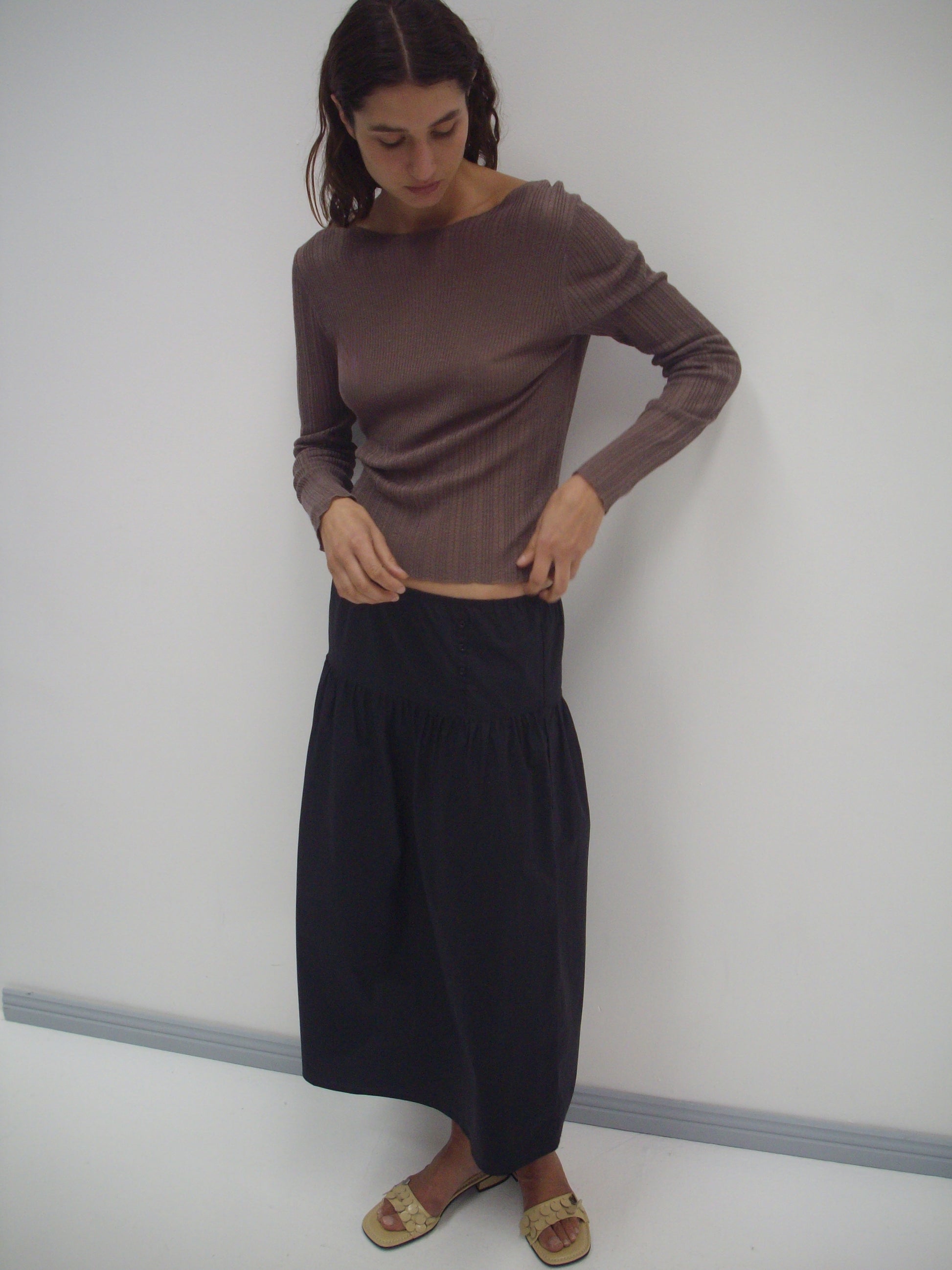 A person with long hair, wearing a fitted brown ribbed long-sleeve top, the Button Skirt - Black, and beige sandals stands against a plain white wall looking down.