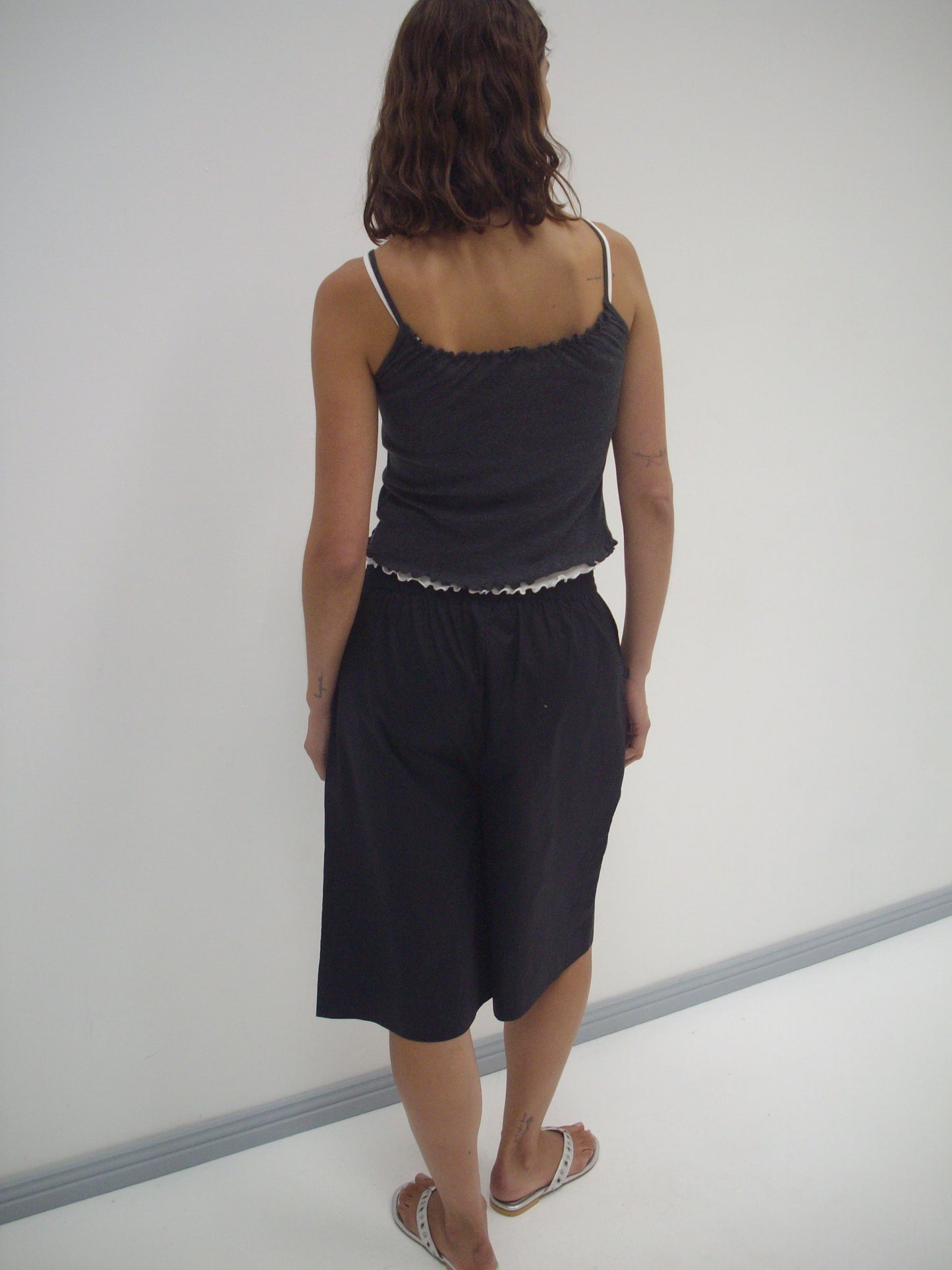 A person with wavy brown hair, wearing the Deiji Studios Long Lounger - Black and flip-flops, stands with their back to the camera facing a white wall.