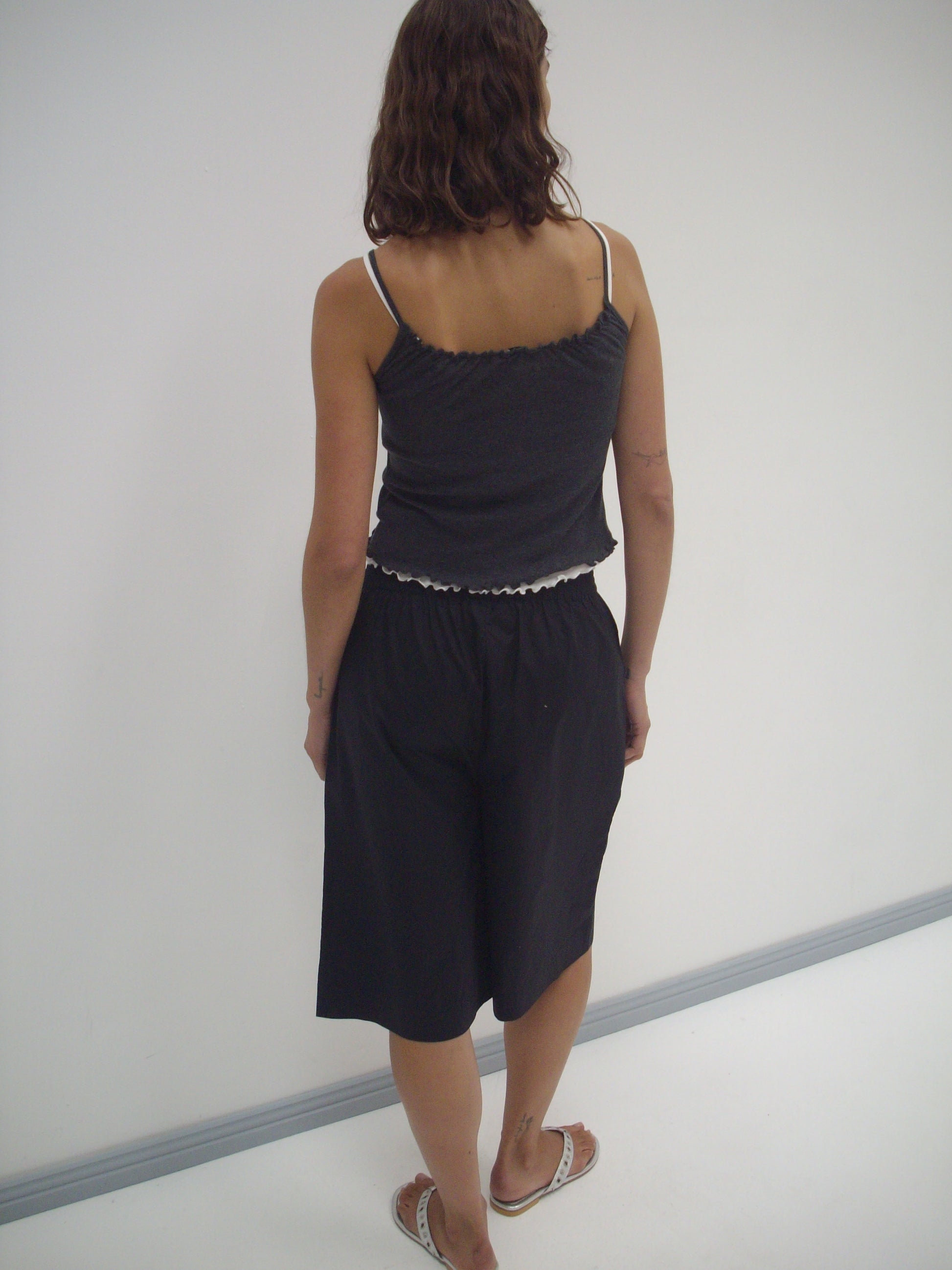 A person with wavy brown hair, wearing the Deiji Studios Long Lounger - Black and flip-flops, stands with their back to the camera facing a white wall.