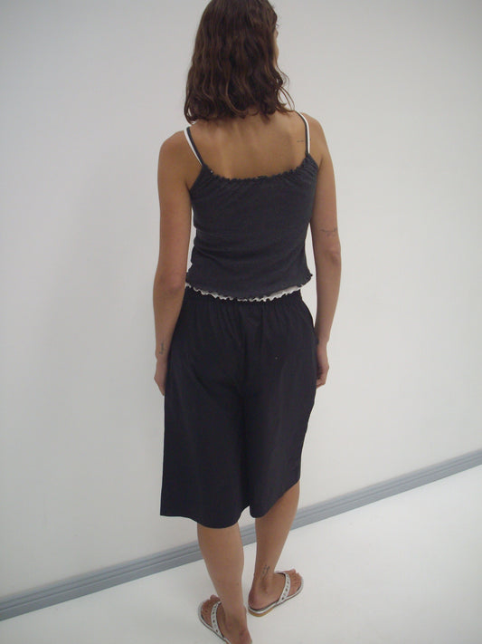 A person with wavy brown hair, wearing the Deiji Studios Long Lounger - Black and flip-flops, stands with their back to the camera facing a white wall.