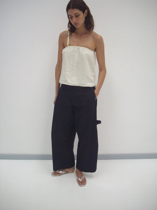 A woman with dark hair stands against a white wall, wearing the Button Strap Top in Pastel Pistachio, loose black wide-leg pants, and silver flip-flops. She looks down with her hands in her pockets.