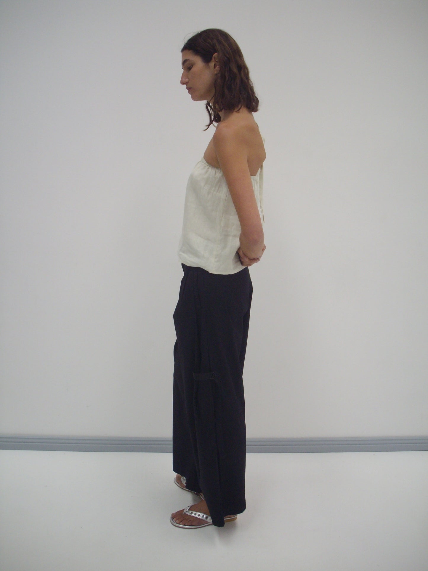 A person with shoulder-length brown hair stands in profile against a white background, wearing the Button Strap Top in Pastel Pistachio, loose black pants, and white sandals. Their hands are behind their back.