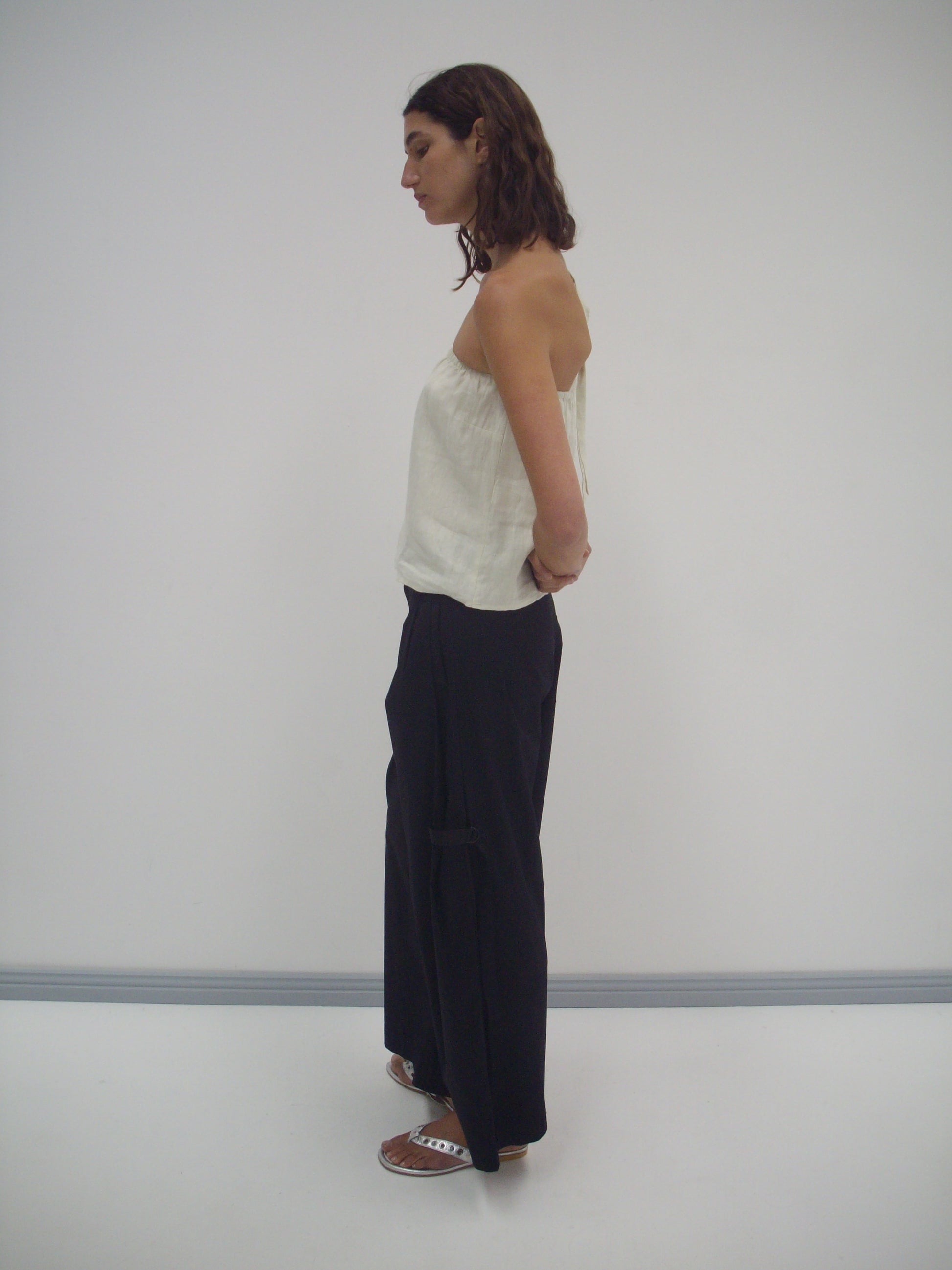 A person with shoulder-length brown hair stands in profile against a white background, wearing the Button Strap Top in Pastel Pistachio, loose black pants, and white sandals. Their hands are behind their back.