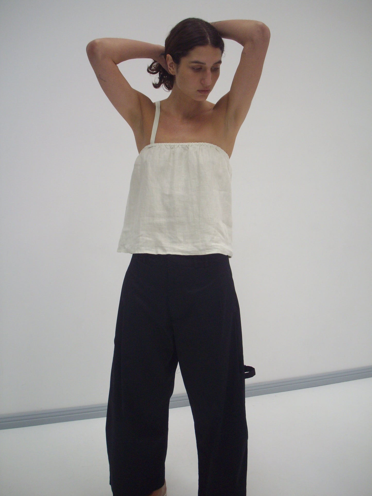 A person wears a Button Strap Top in Pastel Pistachio and loose black pants, standing against a plain white background with arms raised behind their head, looking down thoughtfully.