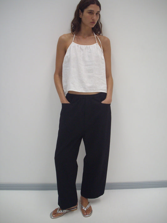 A person with long dark hair stands against a white wall, wearing the Square Pocket Pant - Black (relaxed-fit, organic cotton), a sleeveless white halter top, and sandals. Their hands are in their pockets.