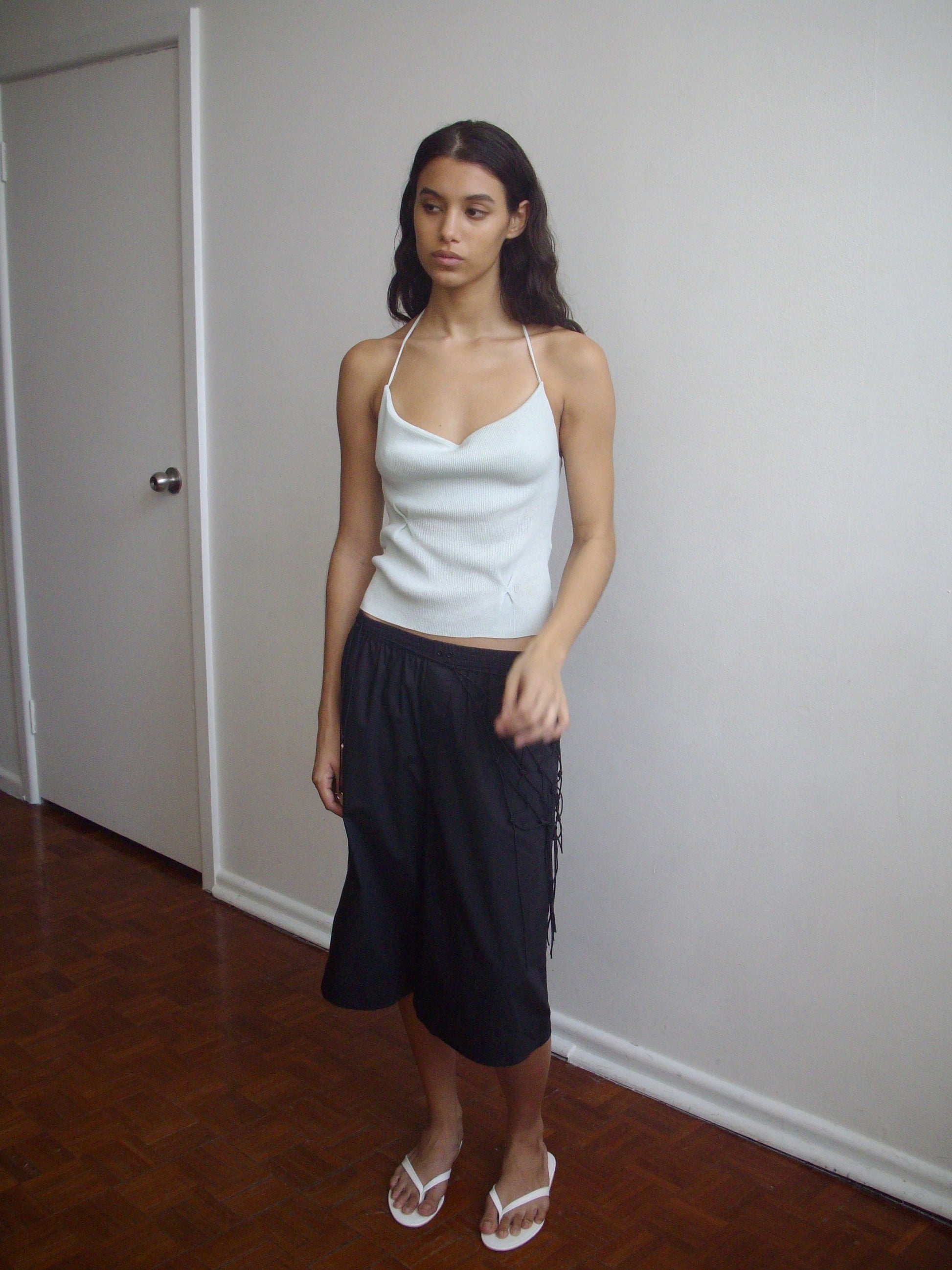 A woman stands indoors against a plain wall, wearing the Long Lounger - Black from Deiji Studios with a light blue camisole and white sandals. She has long dark hair and a neutral expression.