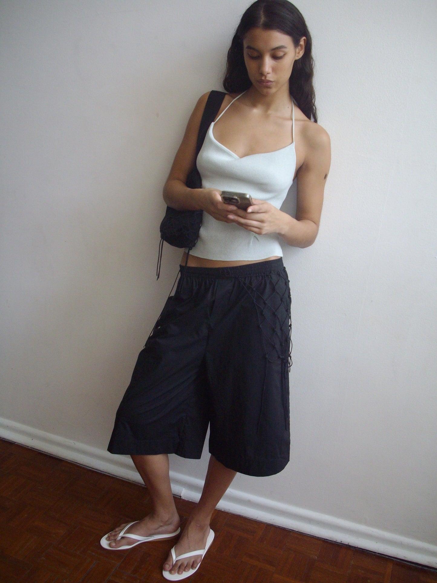 A person stands indoors against a white wall, wearing the Long Lounger - Black, a white organic cotton halter top, white flip-flops, and holding a phone. They have long dark hair and a black bag over one shoulder.