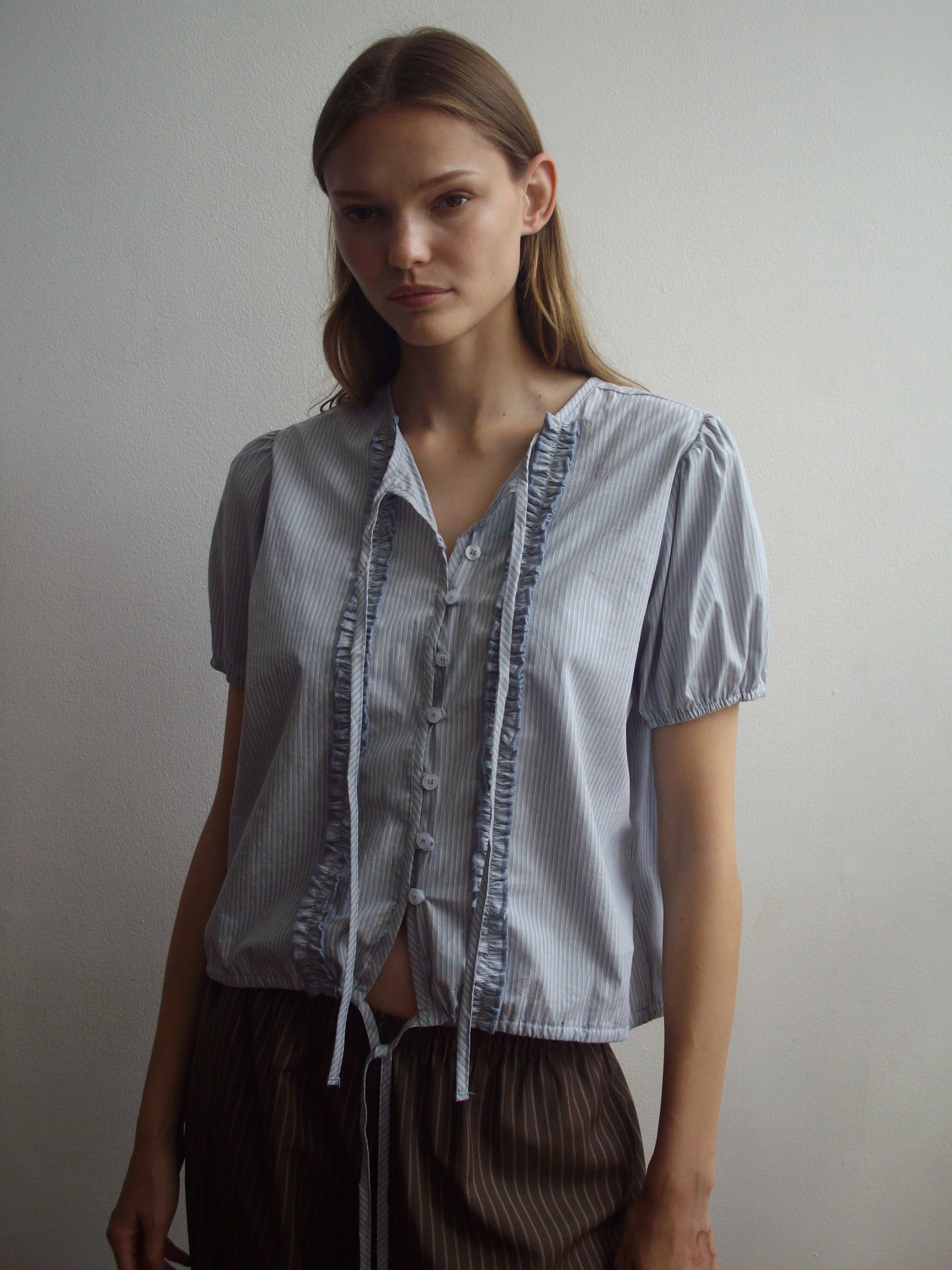 A woman stands against a white wall, wearing the Deiji Studios Double Frill Top - Sky Stripe with ruffle details and ties, paired with dark loose pants. She has long straight hair and a neutral expression.