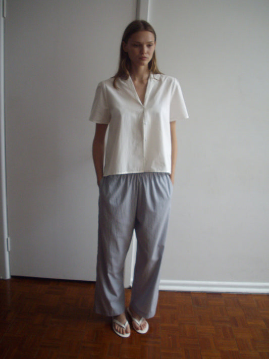 A person stands indoors against a white wall, wearing a short-sleeved white shirt, the Ease Trouser - Glacier Stripe by Deiji Studios, and white flip-flops, with hands in their pockets.