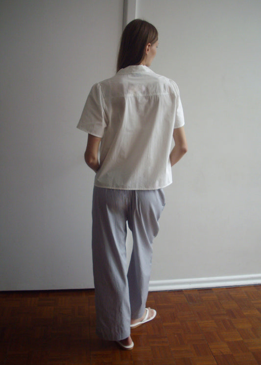 A person stands indoors on a wooden floor beside a white wall, wearing a short-sleeved white shirt and the Ease Trouser - Glacier Stripe by Deiji Studios, paired with white sandals.