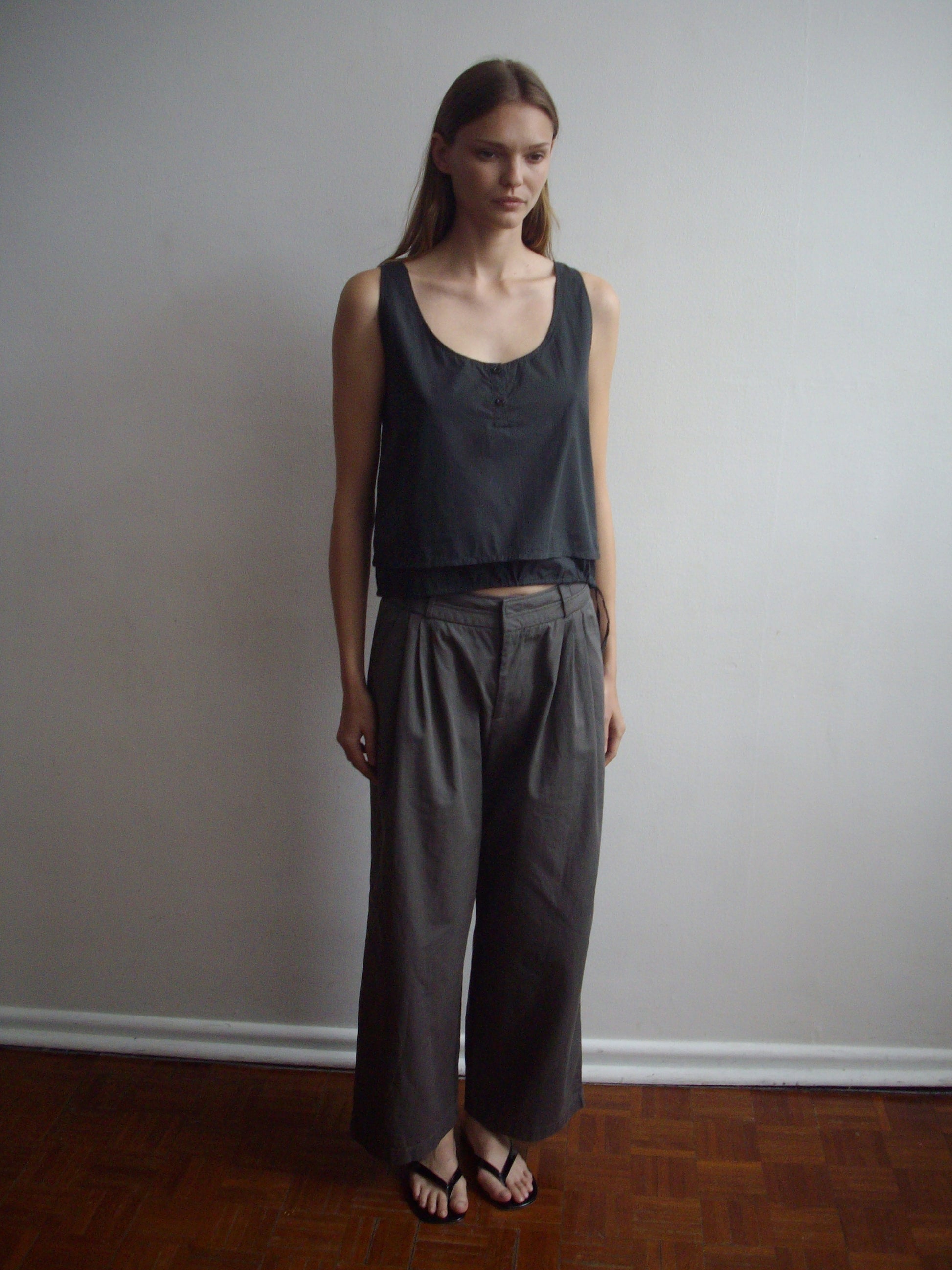 A woman with long straight hair stands indoors against a plain wall, wearing the Layered Top - Black Stripe, loose gray trousers, and black sandals. She has a neutral expression and a relaxed posture.