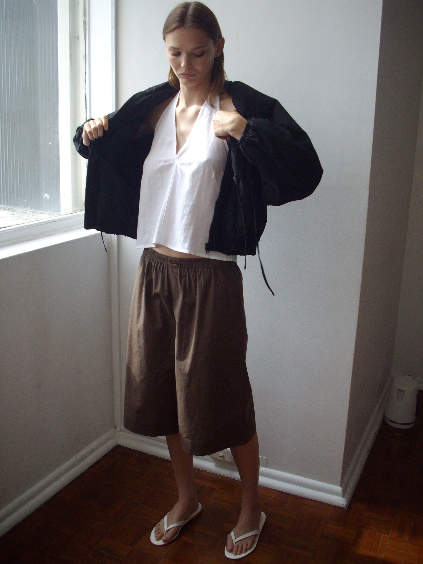 A person stands indoors in sunlight, wearing a black jacket, white sleeveless top, brown knee-length shorts, and white flip-flops—showcasing effortless style inspired by the Long Lounger - Carob Check from Deiji Studios.