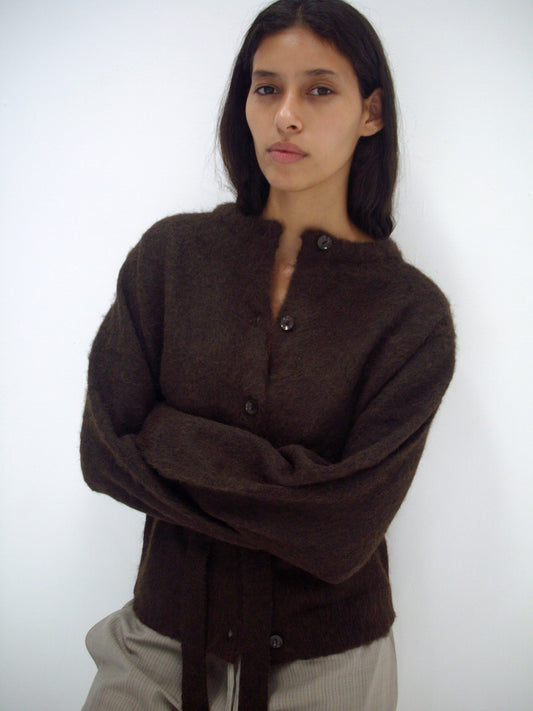 A person with long dark hair stands against a plain white background, wearing the Belted Cardi - Brown from Deiji Studios and light-colored pants, arms crossed with a neutral expression.