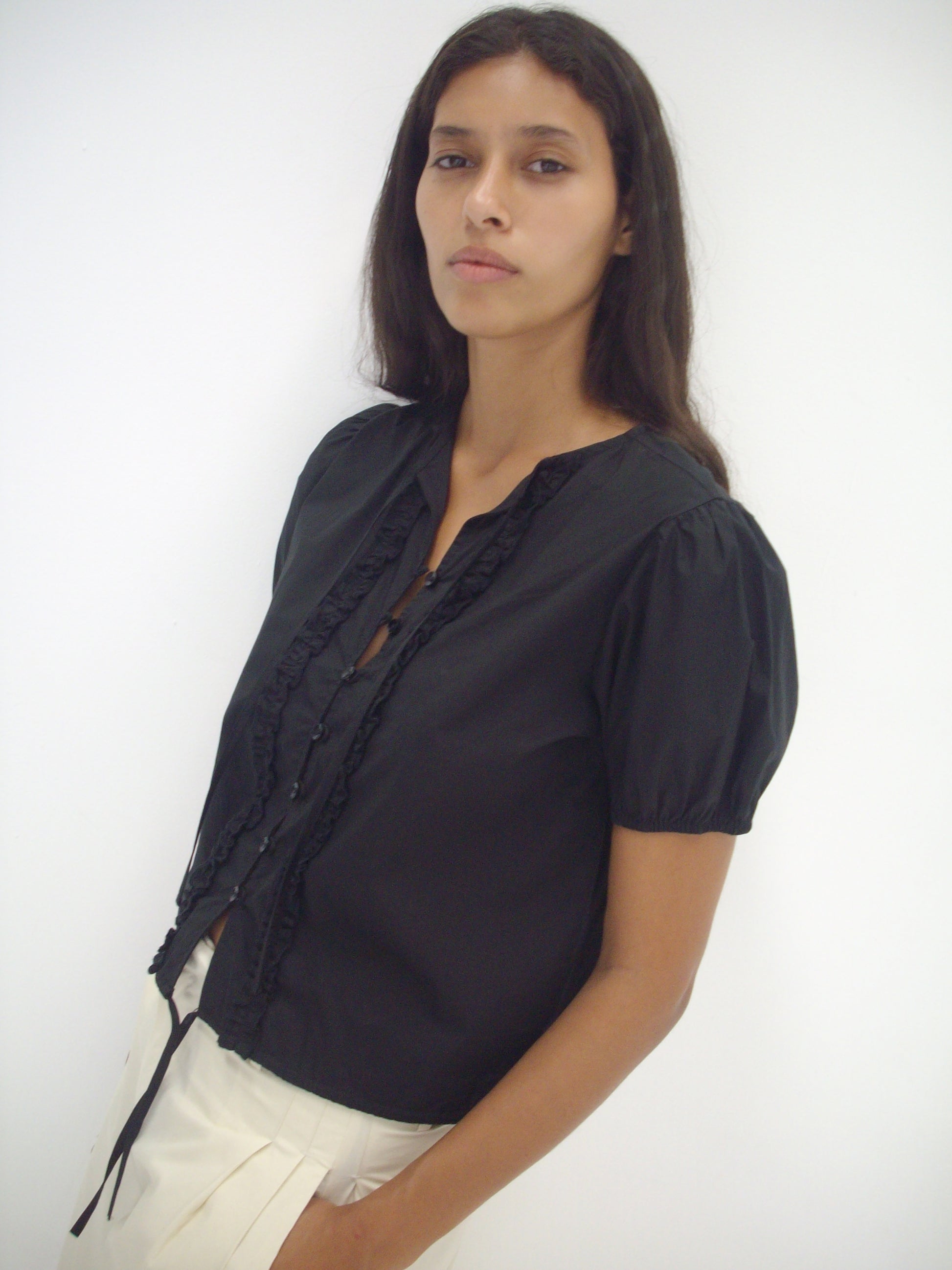 A woman with long dark hair wears the Double Frill Top - Black by Deiji Studios with white pants, standing against a plain white background and looking at the camera with a neutral expression.
