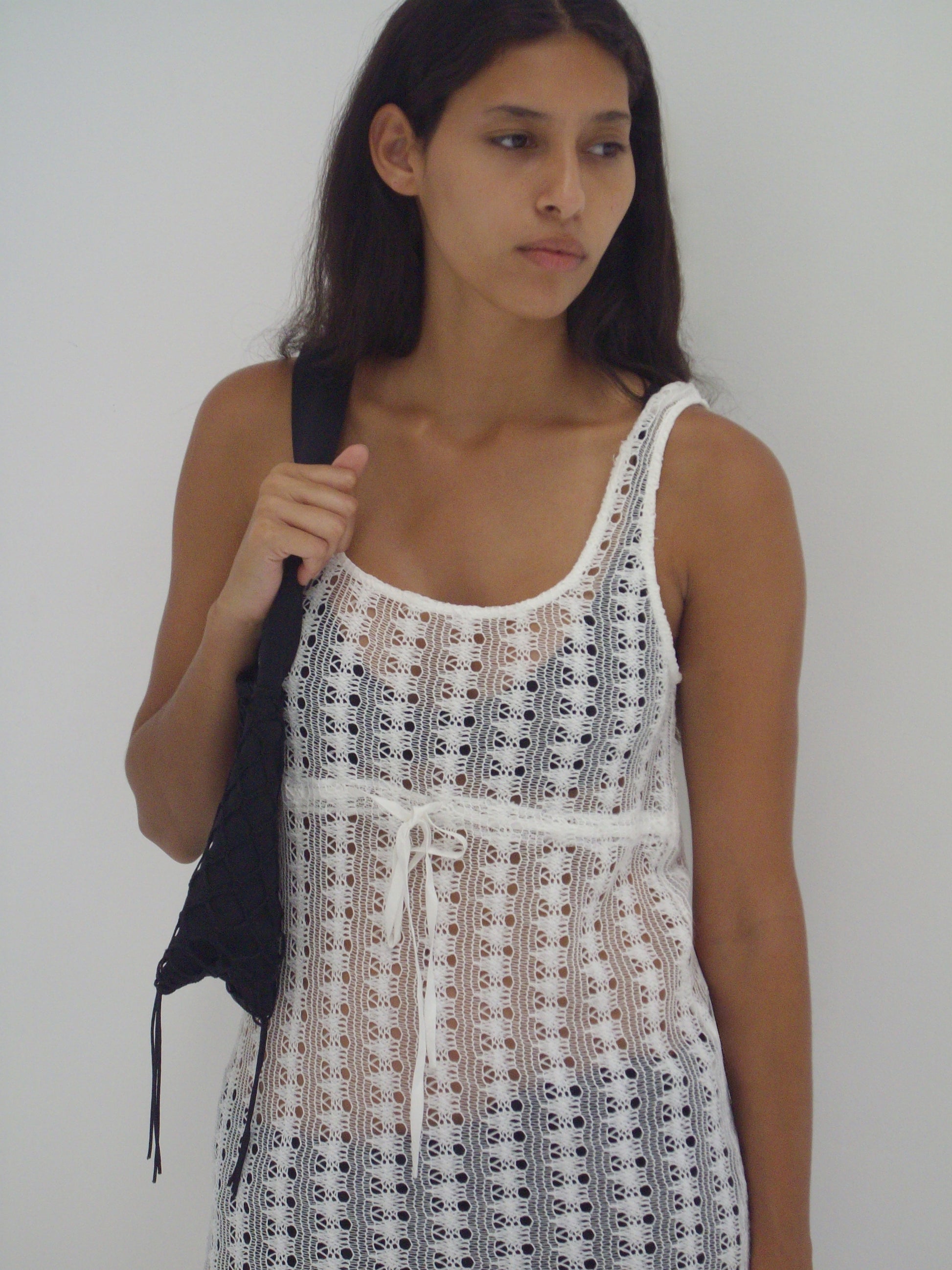 A woman with long dark hair wears the Thread Dress - Off White from Deiji Studios and carries a black woven bag over her shoulder, standing against a plain white background and gazing to the side with a neutral expression.