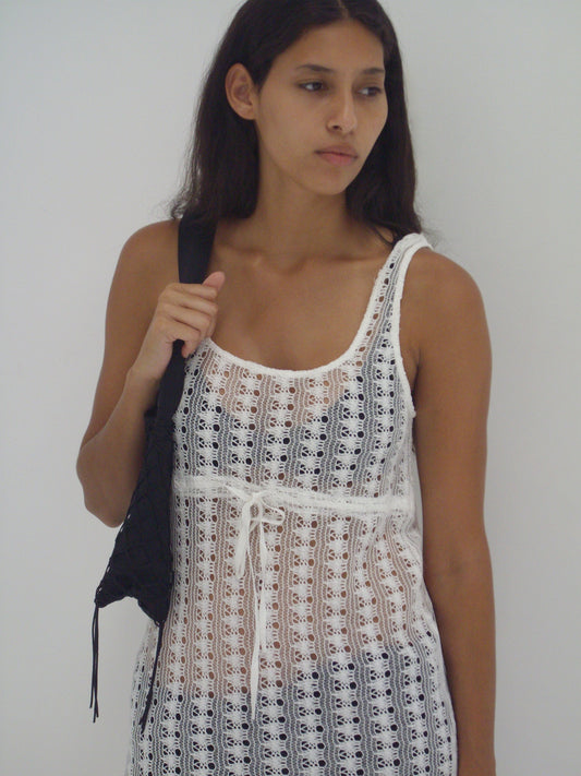 A woman with long dark hair wears the Thread Dress - Off White from Deiji Studios and carries a black woven bag over her shoulder, standing against a plain white background and gazing to the side with a neutral expression.