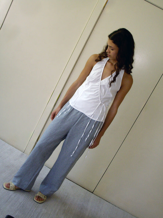 A person with long dark hair stands indoors against a plain light background, wearing a sleeveless white top, sandals, and the linen ease trouser - field check: loose blue-striped French linen pants with an elastic waist.