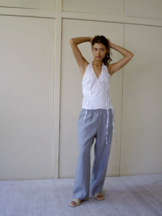 A person stands indoors with arms raised, wearing a white sleeveless wrap top and the linen ease trouser - field check, paired with sandals, in front of cream-colored paneled walls.