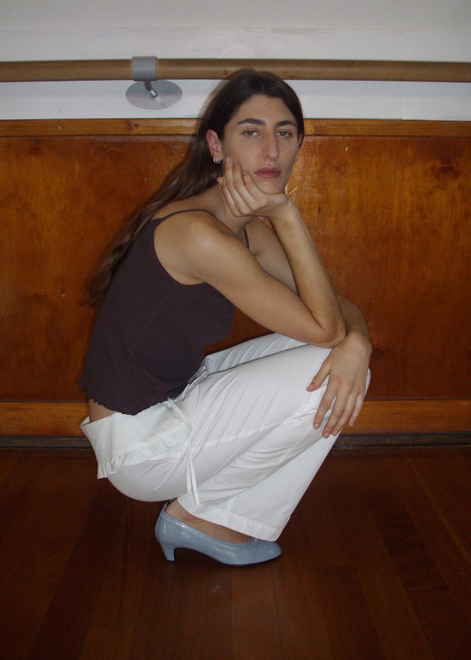 A person with long brown hair, in a sleeveless dark top, Flounce Belt Pant - White from Deiji Studios, and light blue heels squats on a wooden floor with one hand on their chin before a wooden wall and ballet barre.