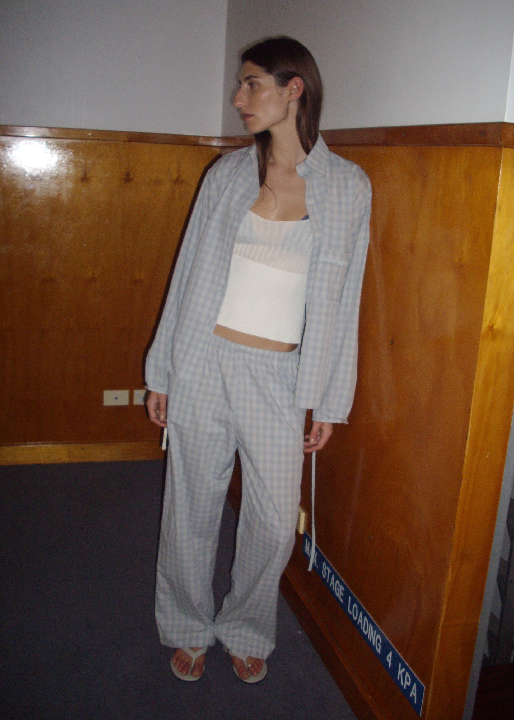 A woman stands indoors in the Side Tie Pant - Blue Check over a white top and sandals, looking to the side near wooden paneled walls and a blue sign on the floor.