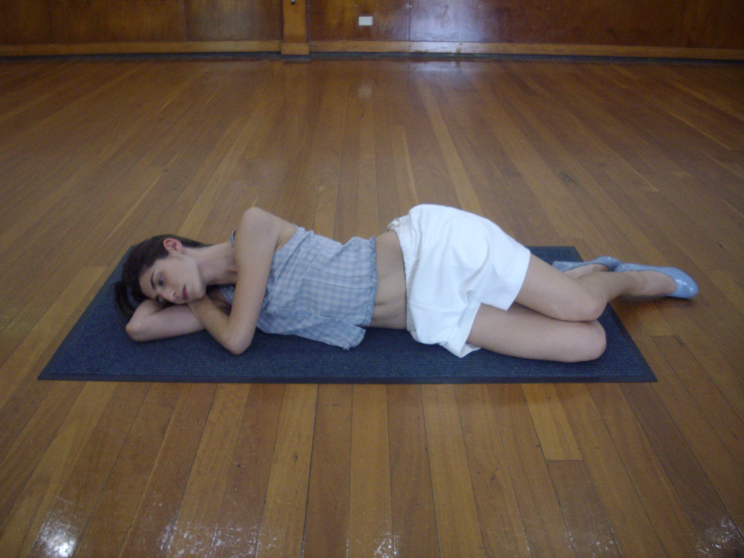 A person lies on their side on a yoga mat in a spacious room, wearing Deiji Studios’ Relax Short - Off White, a sleeveless top, and gray flat shoes.