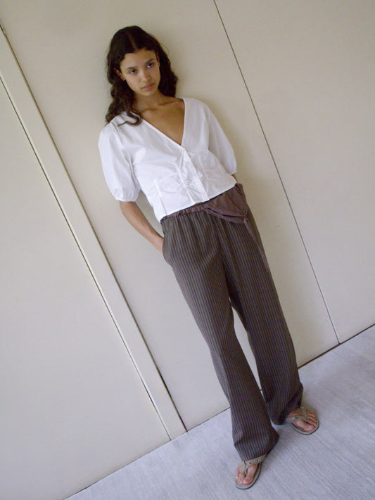 A person stands indoors against a beige wall, wearing the Draw Top - White by Deiji Studios in organic cotton poplin, paired with loose brown pinstriped pants and sandals. They have long, wavy hair and their hands in their pockets.
