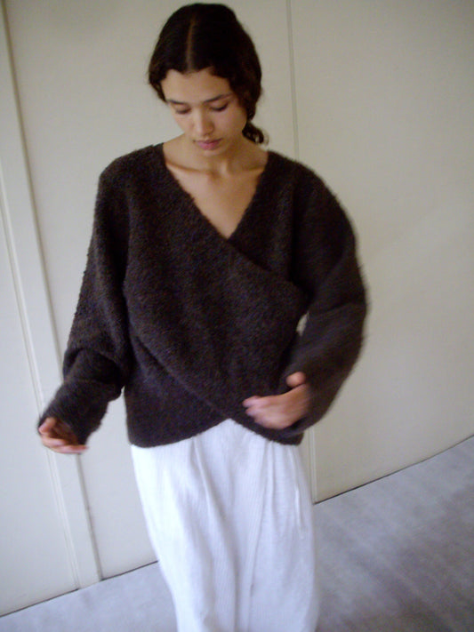 A person with long dark hair wears the Crossover Top - Brown Multi, a sustainable knit from Deiji Studios, paired with white pants. They stand indoors against a plain background, looking down with arms slightly extended.