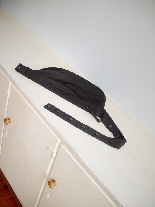 A Press Stud Saddle Bag in dark chocolate recycled polyester sits atop a white cabinet with wooden knobs, set against a plain light gray wall.