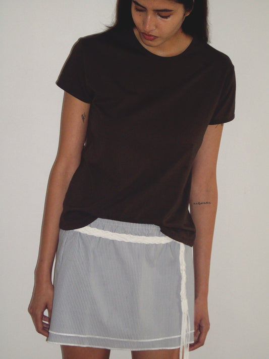 Against a plain background, a person with long dark hair looks down, styled in a black short-sleeve tee and light gray skirt, accessorized with the 0 Plaited Belt - White for adjustable styling.