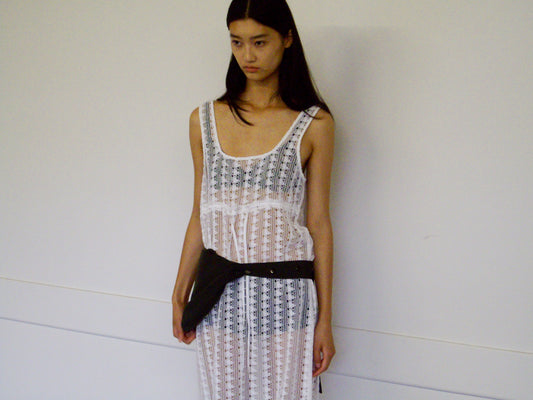 A woman with long dark hair stands against a plain wall, wearing the Thread Dress - Off White by 0 over a black undergarment and a black waist bag. She has a neutral expression, arms by her sides.