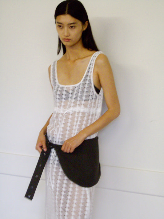 A woman with long dark hair wears a sheer white sleeveless dress, standing against a white wall and holding the Press Stud Saddle Bag in Dark Chocolate.