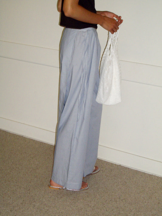 A person stands on a carpeted floor against a plain wall, wearing light blue striped wide-leg pants, a black top, sandals, and holding the Thread Dust Bag - Off White as a shoulder bag.