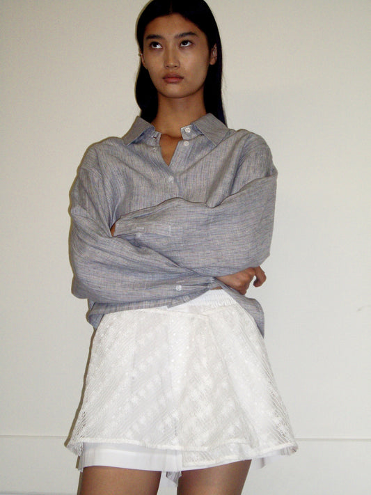 A person with long dark hair stands against a plain wall, wearing a loose gray button-up shirt and the Thread Button Belt - Off White as a short textured skirt. Their arms are crossed and their expression is neutral.