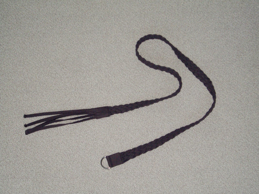 A dark braided leather belt, the Plaited Belt - Chocolate by 0, with fringe on one end and a metal ring on the other, is displayed on a light-colored textured surface.