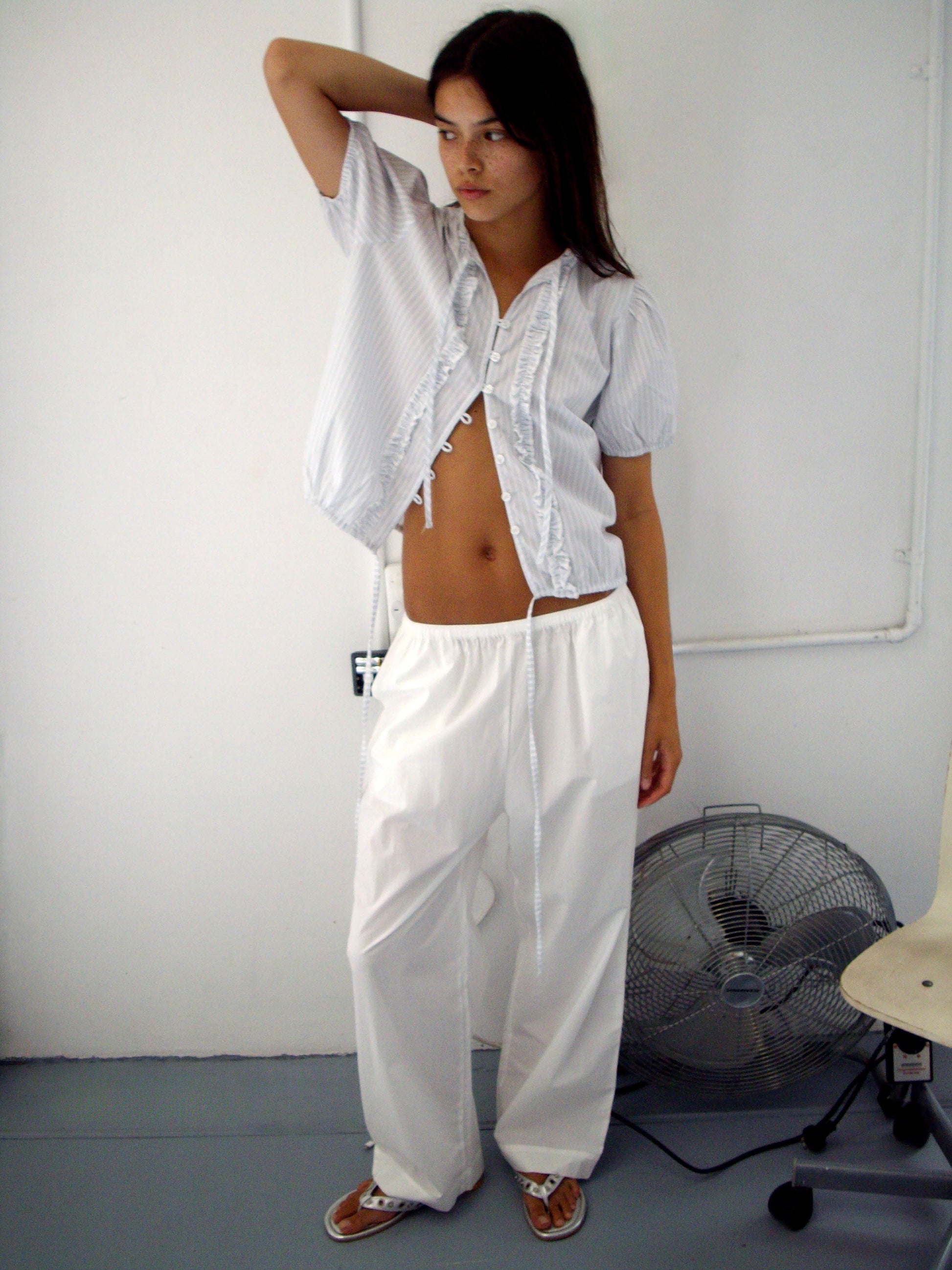 A woman poses indoors in a loose, short-sleeved Double Frill Top - Sage Stripe, partially unbuttoned, paired with baggy white pants. One hand is behind her head; a floor fan and a white chair are in the background.