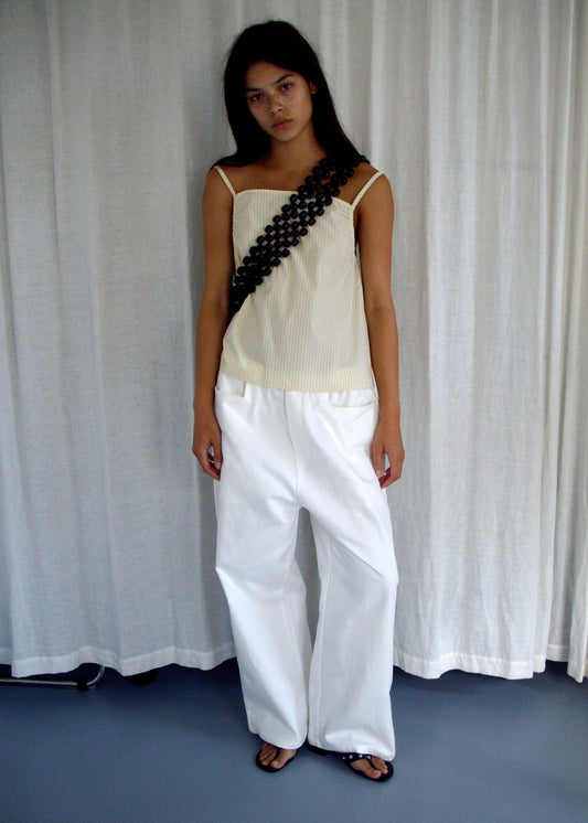 A woman stands before white curtains, wearing the Deiji Studios Gathered Strap Top in Butter Stripe, paired with wide-leg white pants, black sandals, and a statement dark circular link accessory draped across her torso.
