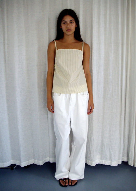 A person stands before white curtains, wearing Deiji Studios' Gathered Strap Top in Butter Stripe (beige, sleeveless, organic cotton) with loose white pants, dark hair down, and black sandals.