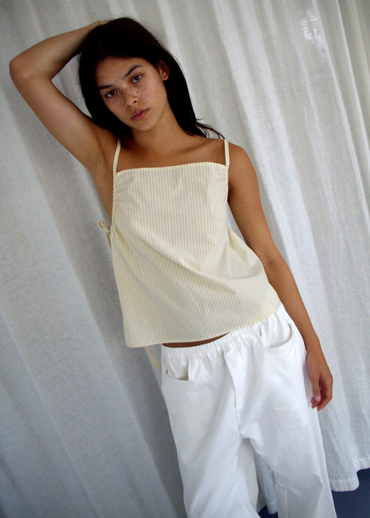 A person with long dark hair poses before white curtains, wearing the Deiji Studios Gathered Strap Top – Butter Stripe in light yellow organic cotton and wide white pants, with their left arm raised and resting on their head.