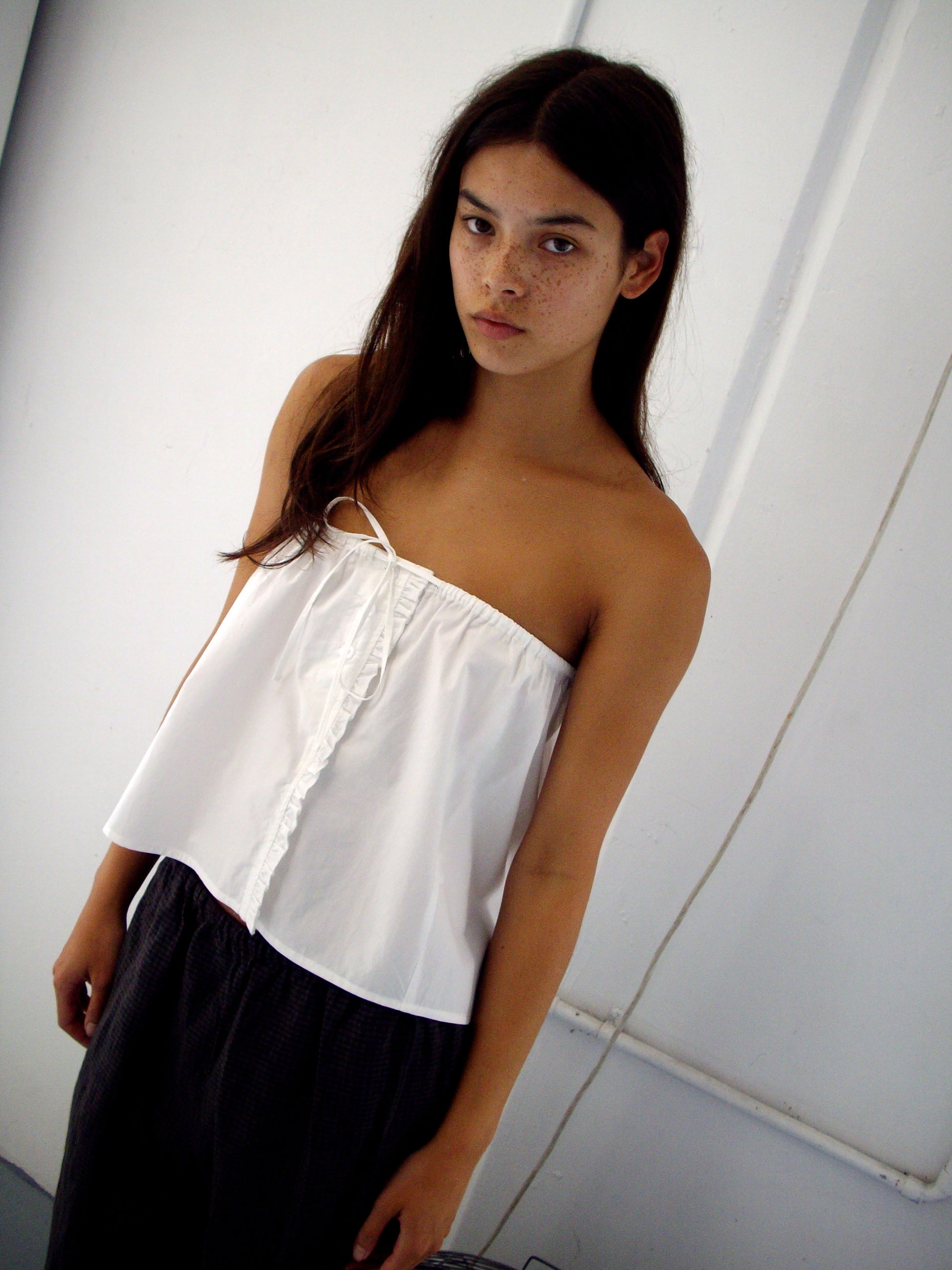 A young woman with long brown hair stands against a white wall, wearing a strapless white top and the Ruffle Mini Skirt - White from Deiji Studios, looking directly at the camera with a neutral expression.