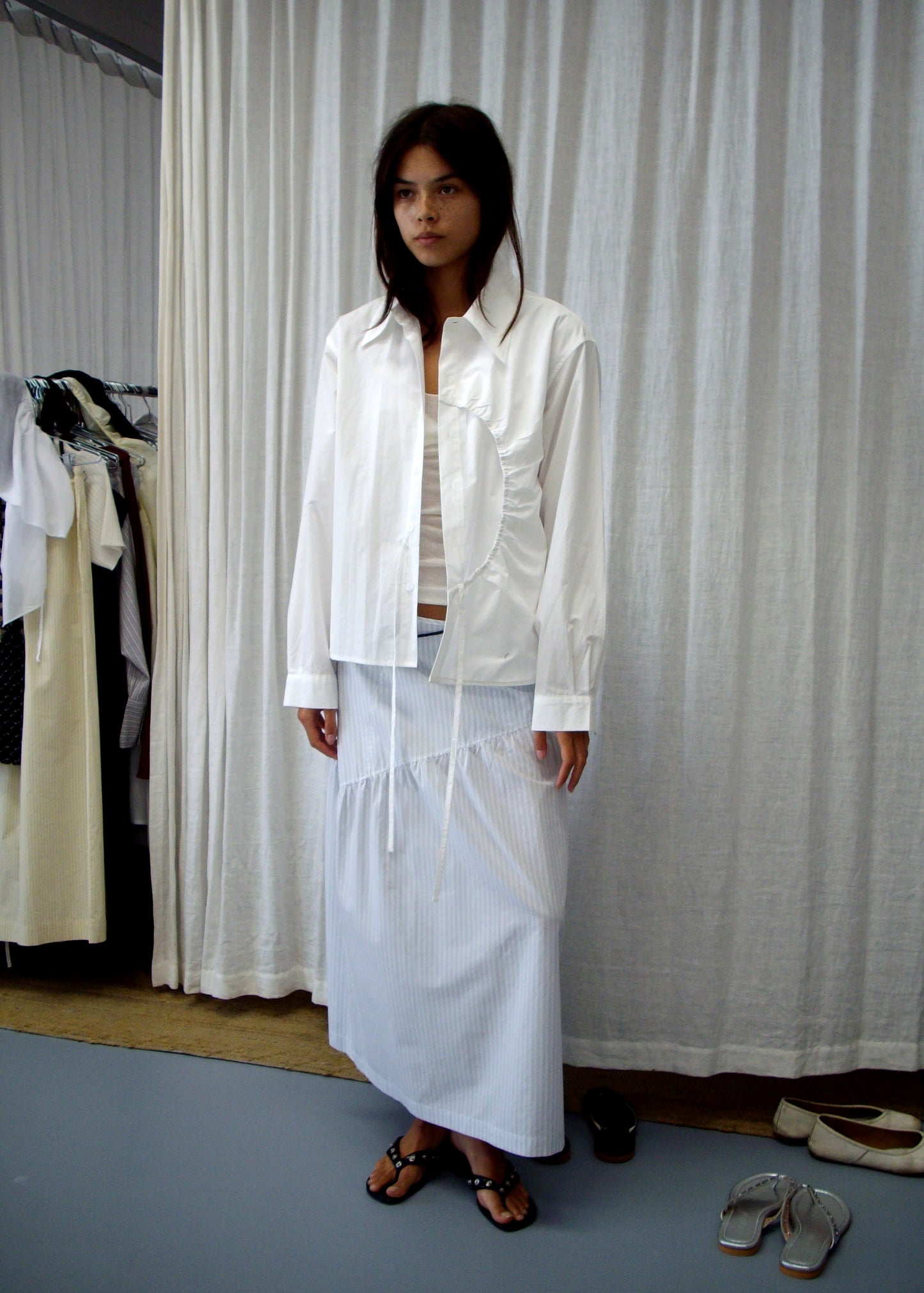 A woman wears the Arch Tie Shirt - White, a loose organic cotton blouse with ruffle details, indoors. She pairs it with a long light blue skirt and black sandals, standing near curtains, clothing racks, and white shoes on the floor.