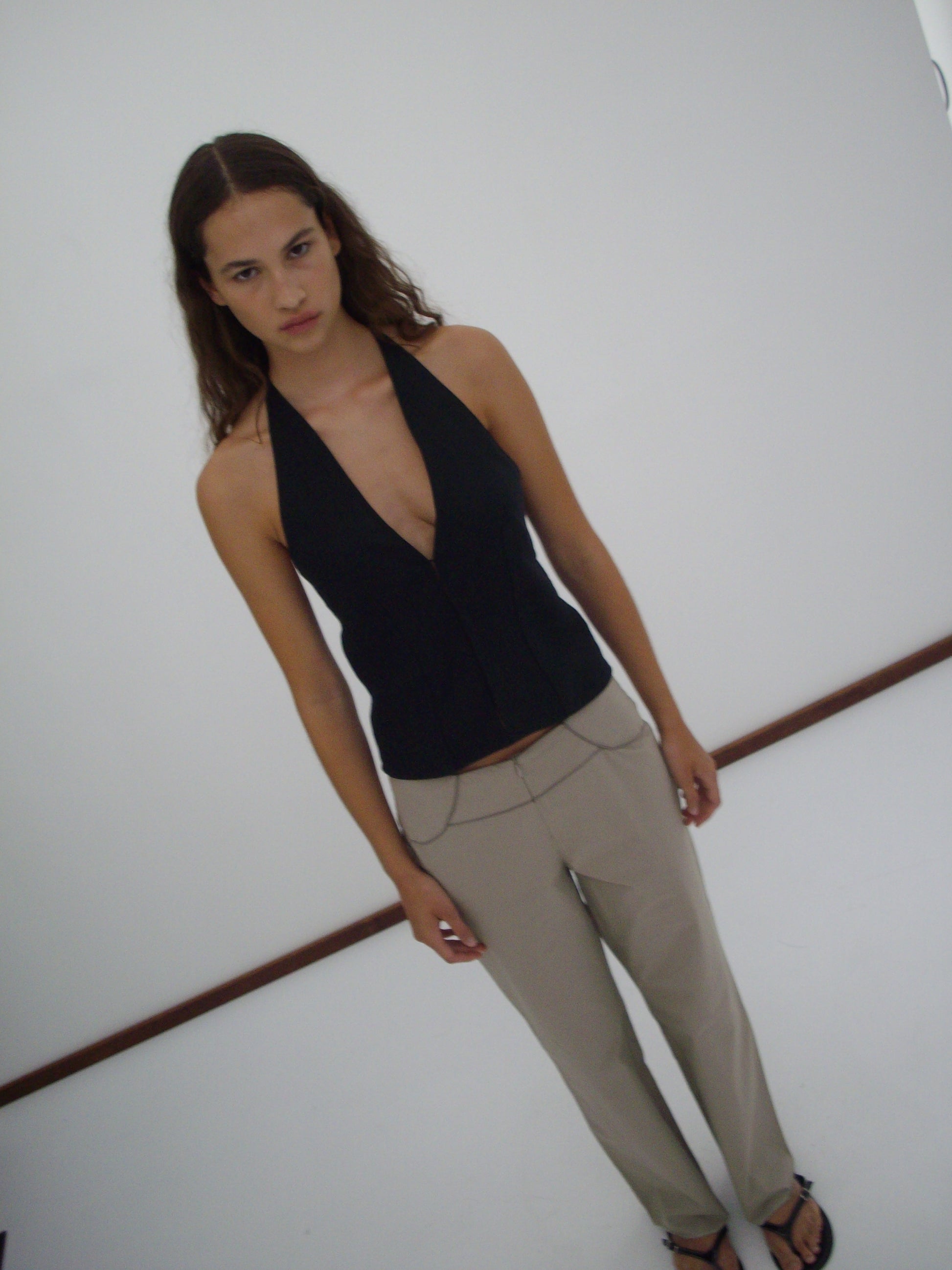 A woman with long, wavy brown hair stands against a plain white wall, wearing the Close Halter Top - Black and beige pants, looking directly at the camera with a neutral expression.