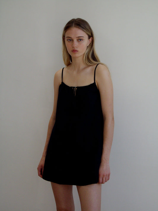 A young woman with long blonde hair stands against a white wall, wearing the Lettuce Mini Dress in black with thin straps. She has a neutral expression and her arms rest relaxed at her sides.